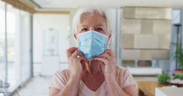 Elderly woman seen putting on a protective face mask in a bright and modern home environment. This can be used to illustrate topics around health and safety during pandemics, elderly care during COVID-19, and hygiene practices in home settings.