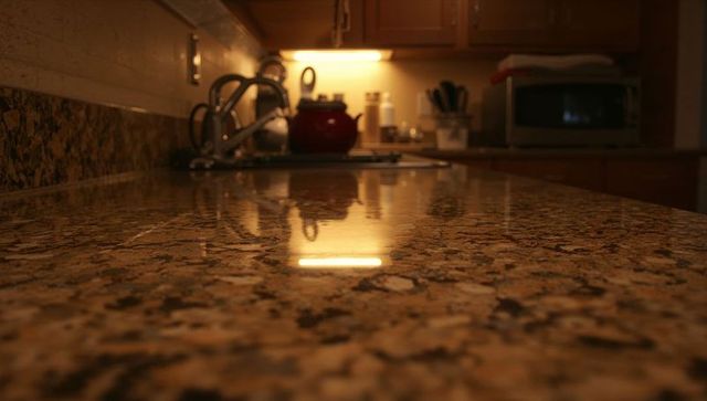 Glossy granite countertop reflecting under-cabinet light with red kettle at sink