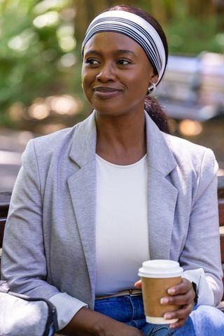 Smiling african american woman holding coffee on park bench wearing gray blazer and headband