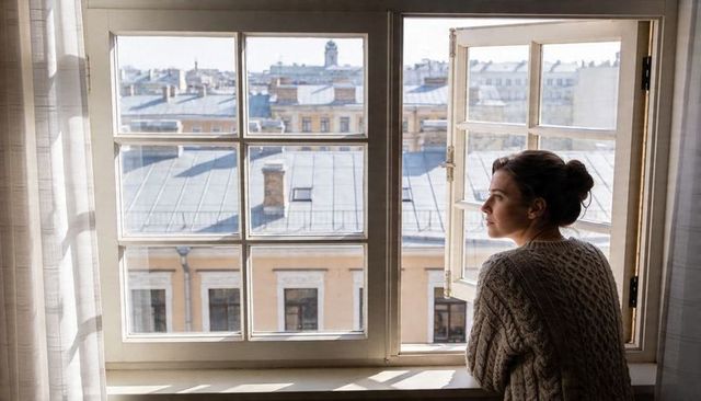 Woman leaning at open apartment window gazing over sunlit city rooftops, contemplative mood