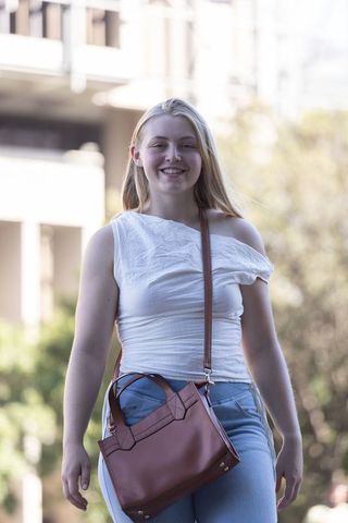 Young woman walking on campus wearing white one-shoulder top with leather crossbody bag