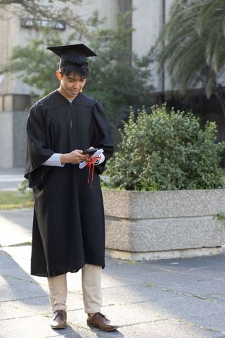 Asian graduate checking phone on campus in cap and gown holding diploma with red ribbon