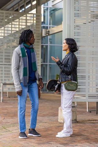 Diverse young adults talking with skateboard and green bag at modern urban plaza