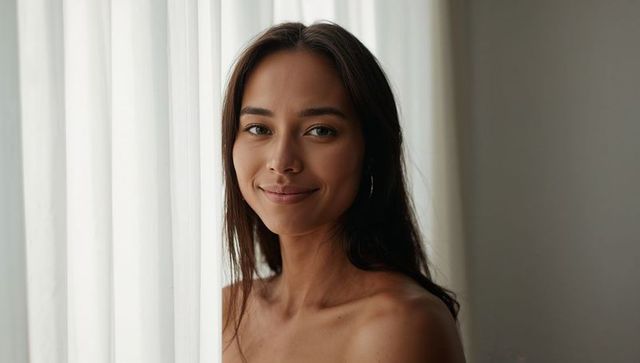 Smiling dark-haired woman posing by sheer curtain in soft natural window light, minimalist headshot