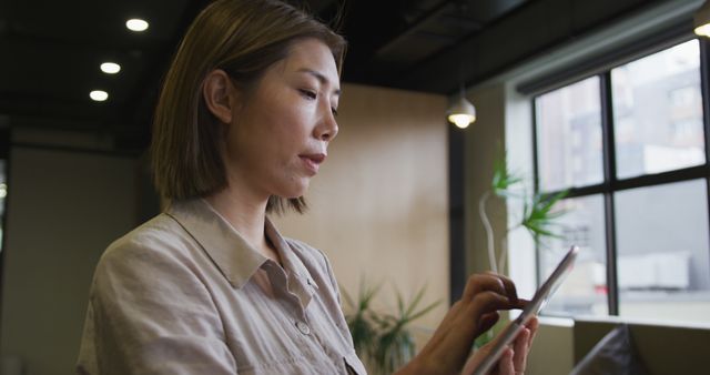 Asian businesswoman standing in modern office using tablet, illustrating a professional working environment. Ideal for articles on modern workspaces, remote work tools, business productivity, corporate settings, technology usage in business, and professional lifestyle blogs.