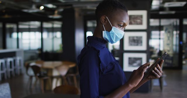 African American businesswoman is using a tablet while wearing a face mask in a modern office environment. Ideal for concepts related to corporate life, remote work, technology integration in business, pandemic safety measures, and workplace health protocols.