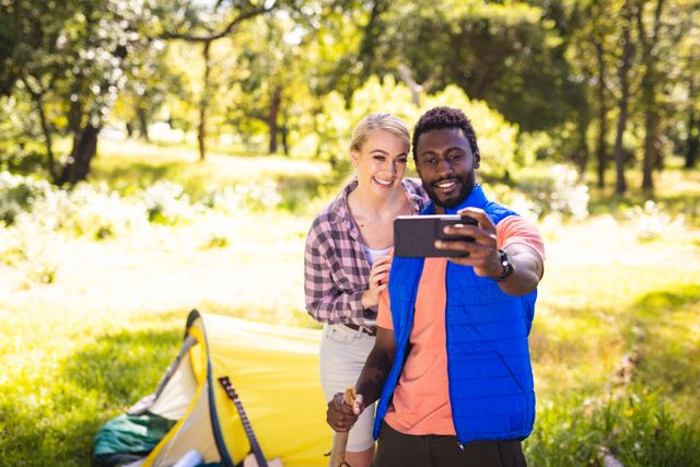Happy diverse couple taking selfie with tent in park. Spending quality time, lifestyle and camping concept.