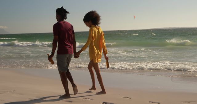 African american couple walking, holding hands and bottles on sunny beach with copy space. Summer, vacations, free time, food and drink, togetherness and lifestyle, unaltered.
