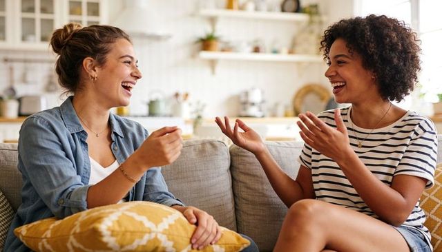Friends laughing together on sunlit living room couch sharing casual conversation