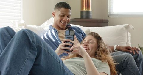 Diverse couple relaxing on light-colored sofa sharing smartphone in sunlit living room