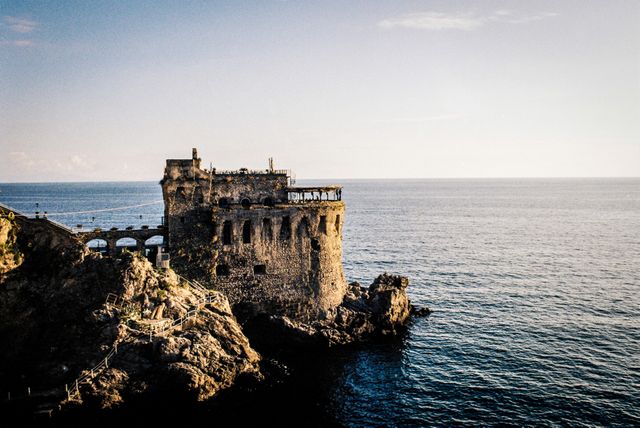 A historic coastal fortress standing on a rocky cliff overlooking the ocean at sunset. This ancient structure exemplifies robust architecture and offers a picturesque view of the sea. Ideal for use in travel promotions, historical site brochures, destination posters, and educational materials on medieval architecture and coastal defense.