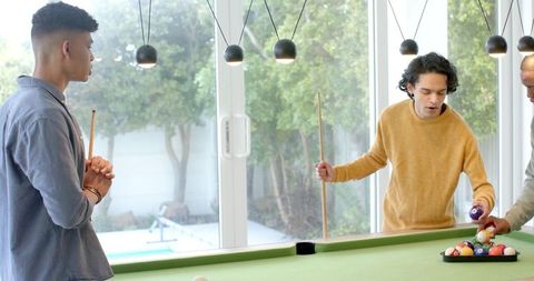 Diverse friends playing pool in sunlit game room with glass doors and backyard view