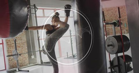 Female athlete executing high roundhouse kick on heavy bag in bright industrial gym
