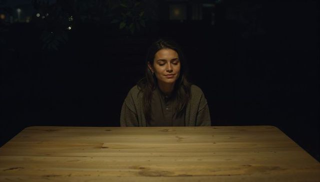 Solitary woman sitting at wooden table closing eyes under warm patio lamp, nighttime calm