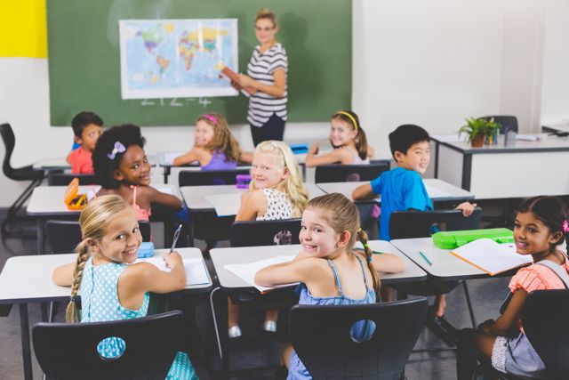 This image shows a diverse group of school kids smiling and looking back at the camera during a geography class. The teacher is pointing at a world map on the board. Ideal for use in educational materials, school brochures, websites promoting primary education, and articles about multicultural classrooms and learning environments.