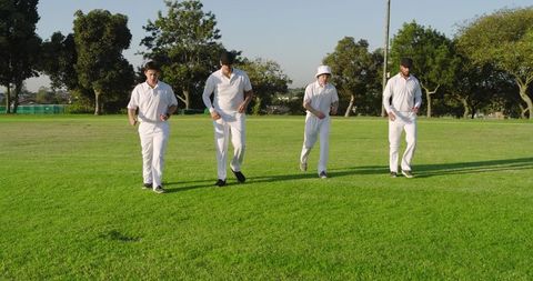 Four cricket players jogging across outfield in white uniforms on sunny green field