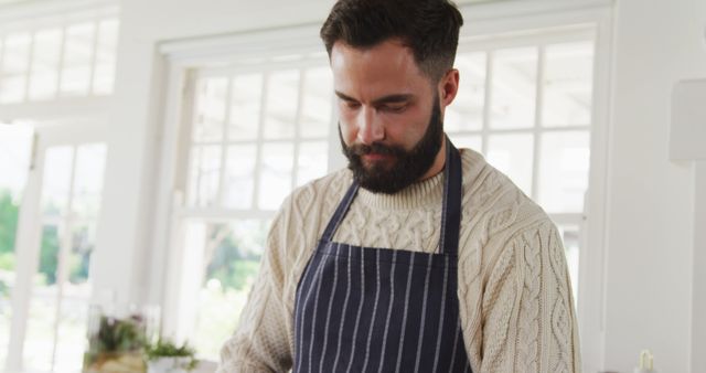 Bearded man wearing striped apron preparing food in bright modern kitchen. Great for content about home cooking, kitchen design, culinary arts, and lifestyle blogs.
