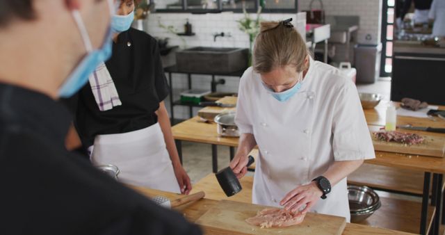 Professional chef wearing a mask is demonstrating meat preparation techniques to students in a commercial kitchen. This image is ideal for depicting culinary education, professional cooking classes, food and hospitality industry training, hygiene practices in the kitchen, or health and safety regulations in culinary arts.