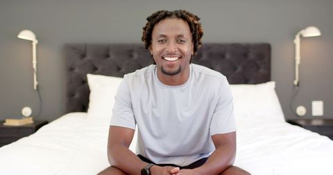Smiling african american man sitting on bed in modern minimalist bedroom with smartwatch