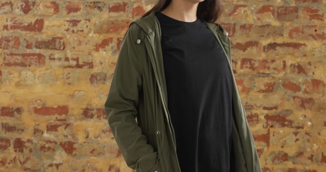 Young woman stands confidently wearing green jacket and black t-shirt, with exposed brick wall background. Useful for lifestyle, fashion, urban living, street style, and casual clothing themes.