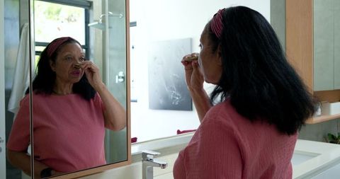Senior african american woman using facial roller at bathroom mirror for skincare