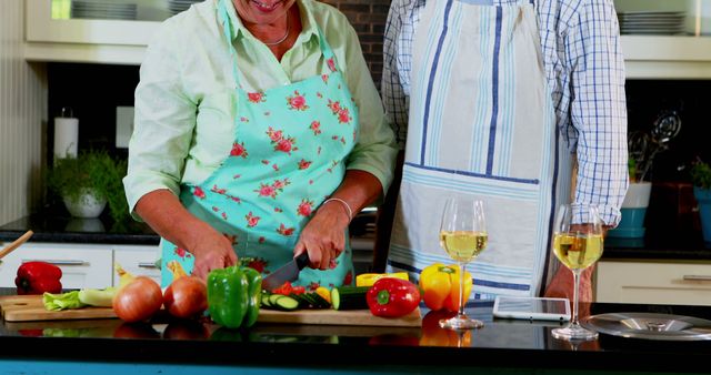 Smiling couple chopping fresh vegetables while preparing a meal in a modern kitchen. The countertop is covered with colourful produce, including bell peppers, onions, and herbs. The couple is wearing aprons and engaging in food preparation, creating a warm and inviting scene. Perfect for use in articles or advertisements about healthy eating, family cooking, modern kitchen designs, or promoting recipes and culinary teamwork.
