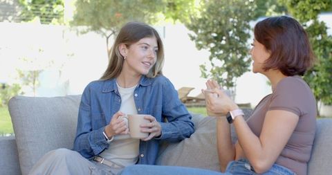Female Friends Chatting Over Coffee on Outdoor Sofa in Sunny Backyard Patio, Cozy Morning