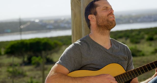The image depicts a man playing an acoustic guitar outdoors, enjoying a serene and natural environment. The expression on his face suggests he is deeply immersed in the music. This image is perfect for use in contexts related to music, leisure activities, outdoor relaxation, and lifestyle promotions.