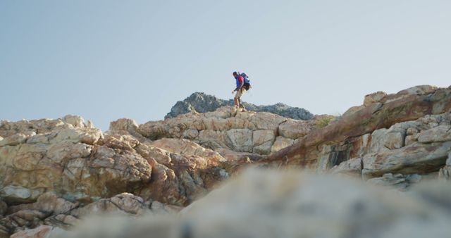 Hiker wearing backpack climbing rocky landscape with clear sky overhead. Ideal for themes related to adventure, outdoor activities, physical fitness, travel, exploration of nature, and mountaineering.