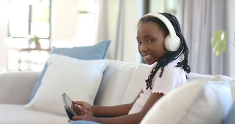 African girl listening to headphones while using tablet on bright sofa at home