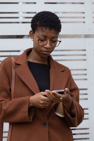 African american woman checking smartphone and holding coffee, brown coat urban commuter
