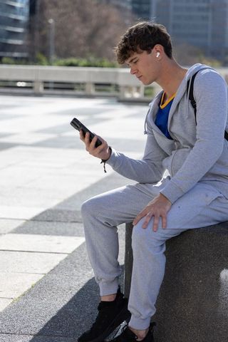 Teen male sitting on concrete bench in modern plaza holding smartphone and listening with earbuds