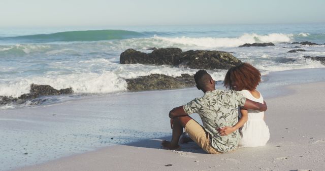 Couple in casual attire sitting on beach sand, enjoying a romantic moment by the ocean. Great for use in vacation promotions, romantic getaways, lifestyle blogs, and social media posts emphasizing love and relaxation.