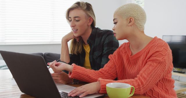 Two young women are collaborating and working on a laptop in a modern home. They are focused and discussing their work while sitting at a wooden table. Could be used for concepts such as teamwork, remote work, productivity, or modern lifestyles.