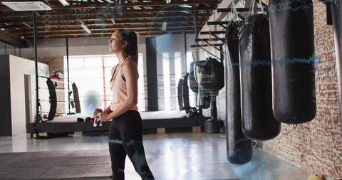Female athlete preparing for boxing workout in loft gym holding jump rope near heavy bags