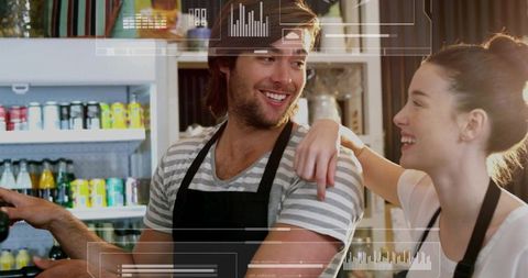 Smiling baristas laughing and serving behind cafe counter with drink fridge and hud