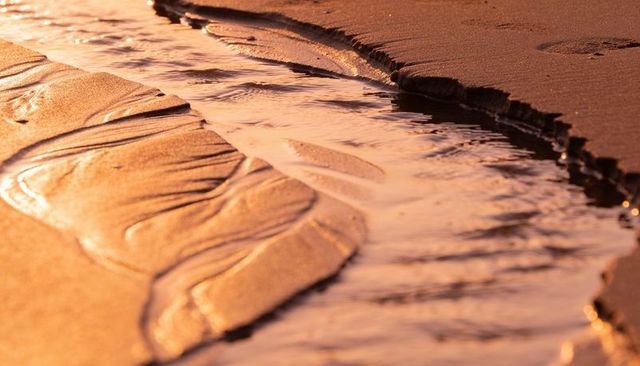 Golden hour tidal channel flowing through wet sand with ripples, footprints and reflections