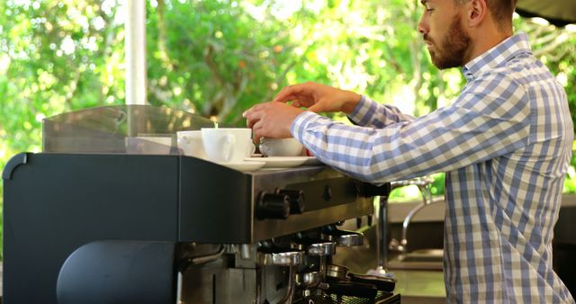 Barista preparing coffee using an espresso machine at an outdoor cafe. The barista wears a plaid shirt, and the background showcases lush greenery, suggesting a relaxed outdoor setting. Ideal for use in content about cafe culture, barista training, coffee brewing tutorials, or casual dining environments.