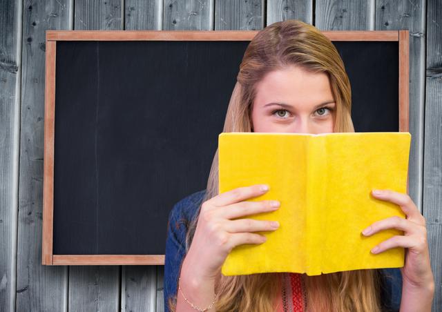 Teenage girl holds bright yellow book in front of blackboard and wooden background. Ideal for educational materials, study websites, or classroom decor. Captures essence of learning, curiosity, and academic environment.