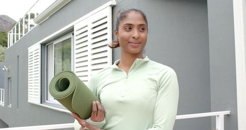 Indian woman holding rolled yoga mat on balcony wearing mint activewear, outdoor wellness