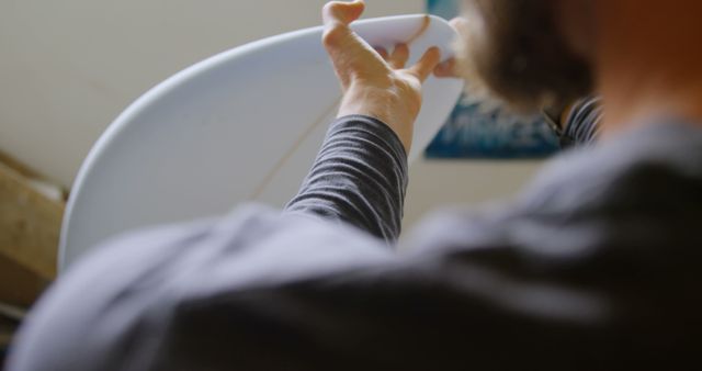 A craftsman is shaping a surfboard with great precision in a workshop. His hands and a part of the surfboard are in focus, reflecting dedication and skill. Perfect for content about handmade surfboards, surf culture, artisanal workshops, or woodworking craftsmanship.