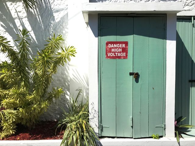 Warning sign on old green door surrounded by tropical plants reflects attention to safety and maintenance. Suited for content emphasizing safety protocols, electrical warnings, industrial settings, or tropical environments.