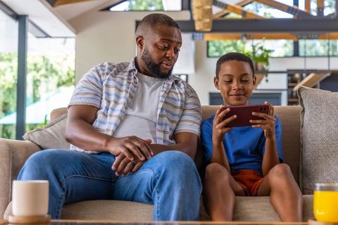 African american father and son sharing smartphone on sofa laughing together in home