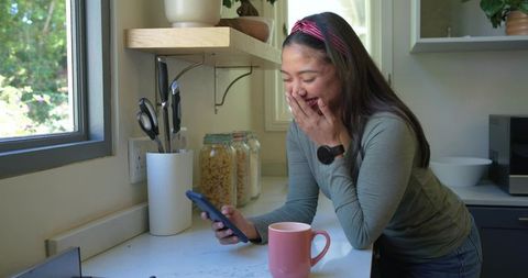 Laughing woman leaning on kitchen counter checking smartphone morning message reaction