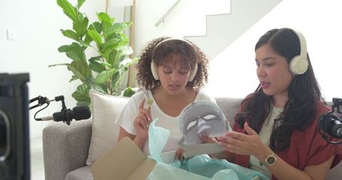 Diverse female hosts unboxing gray led facial mask while filming skincare review on sofa