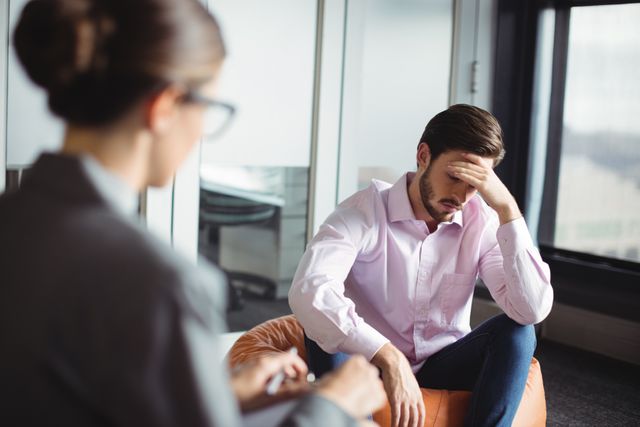 Man consulting with a counselor during a therapy session. The man appears stressed, holding his head in his hand, while the counselor listens attentively. This image can be used for articles or websites related to mental health, therapy, counseling services, emotional support, and professional help.