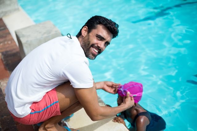 Male instructor assisting a young swimmer with her swim cap at the poolside. Ideal for use in materials promoting swimming lessons, water safety, summer activities, and sports training. Can be used in educational content, advertisements for swimming schools, and articles about mentorship and childhood education.