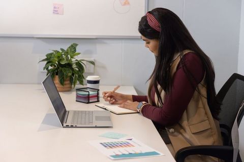 Mid adult woman writing at desk with laptop and charts, focused modern office workspace