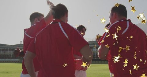Red team huddle celebrating victory with gold confetti on sunlit stadium pitch backlit