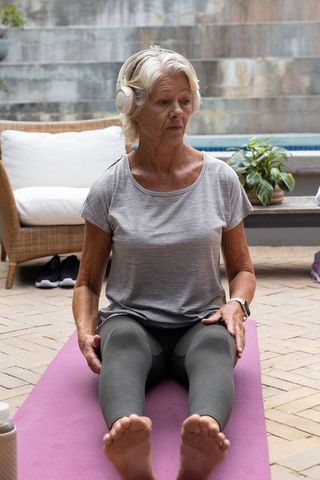 Senior woman practicing seated yoga on patio near pool wearing headphones and smartwatch